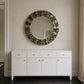 A round wall mirror with a textured, metallic gold and silver mosaic border hangs on a beige wall above a white wooden console cabinet with gold knobs.