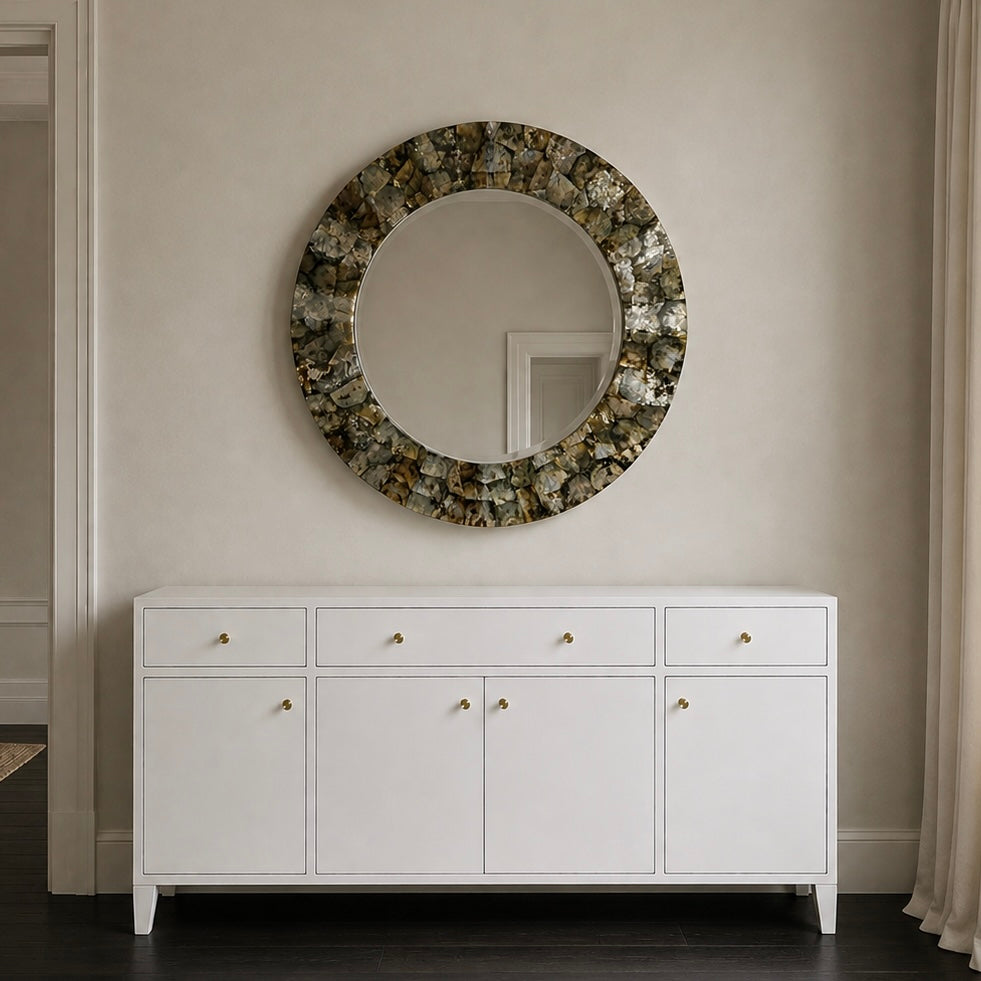 A round wall mirror with a textured, metallic gold and silver mosaic border hangs on a beige wall above a white wooden console cabinet with gold knobs.