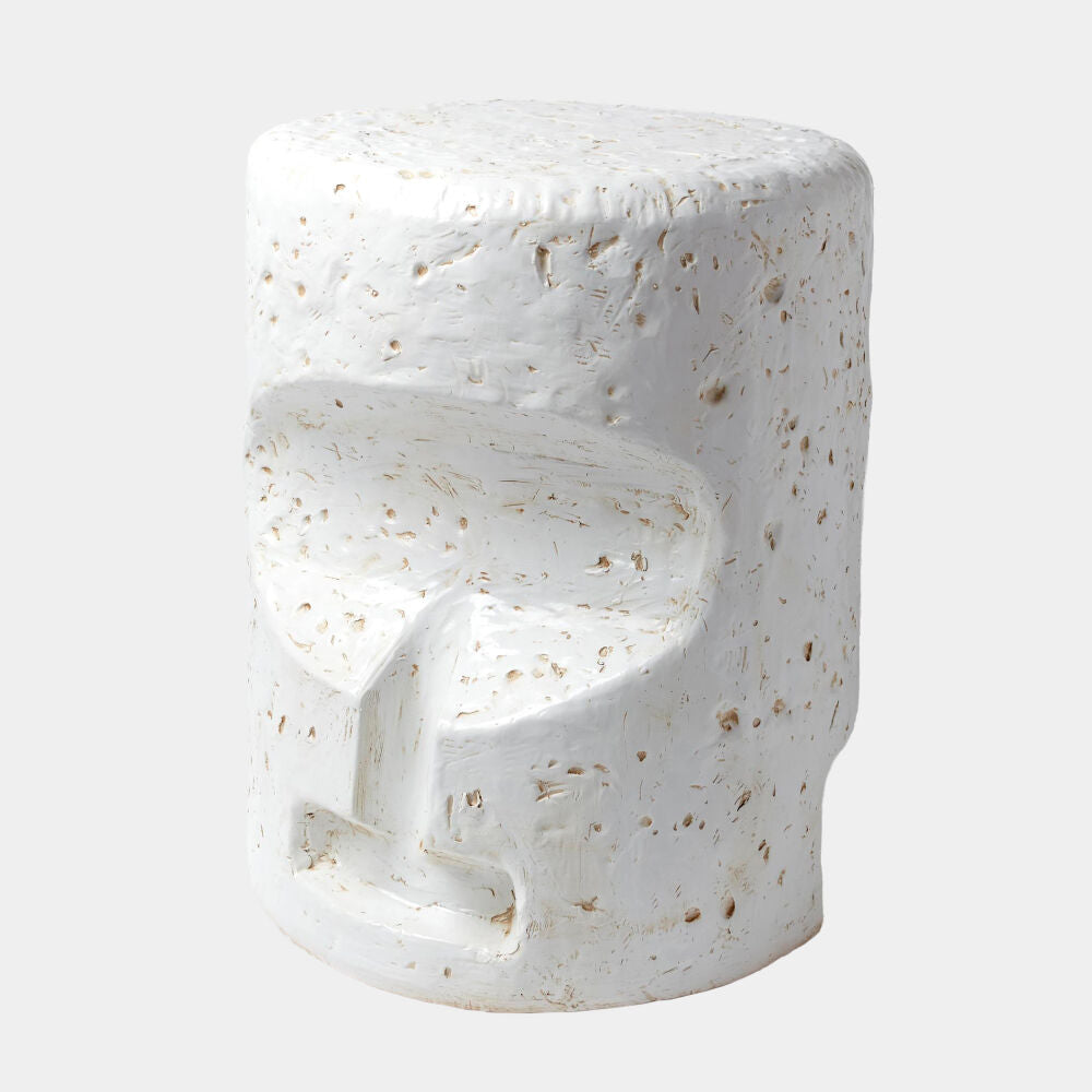 A modern sculptural side stool with a textured, organic shape resembling a stylized face, finished in white with natural beige stone flecks, set against a light gray background.