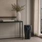 A modern interior scene with a dark textured console table, a white ribbed vase holding dried branches, a rectangular stone tray, and a dark sculptural stool beside a window with sheer curtains.