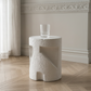 A textured white cylindrical drinks table with an abstract sculptural design, shown in a bright room with a light wood herringbone floor, white walls, and a clear ribbed glass cup on top.