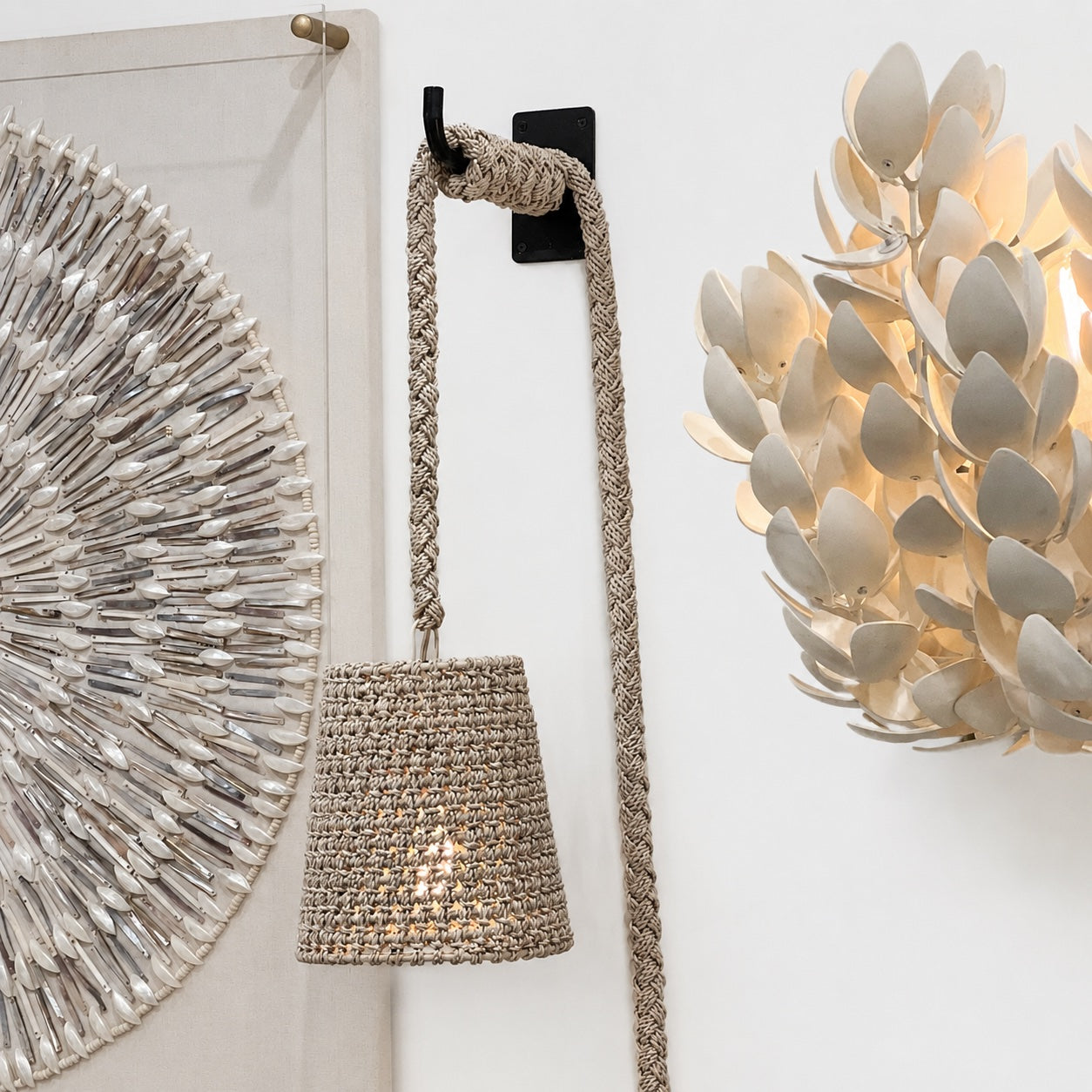 A woven rope pendant light with a conical shade hanging from a black wall hook, shown in a styled interior setting with a decorative wall mirror and a lit leaf-shaped wall sconce.