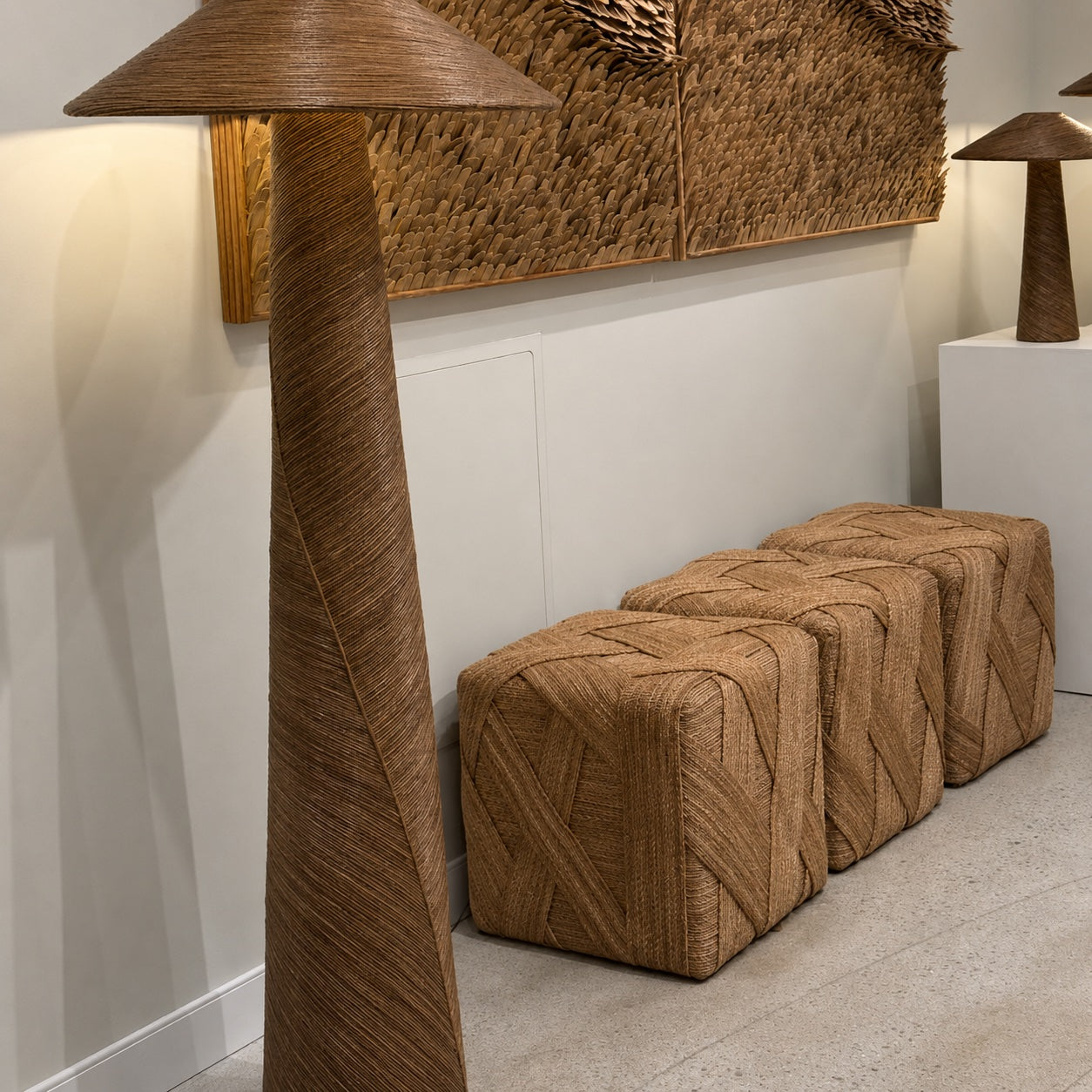 A modern interior scene featuring a tall floor lamp and three woven ottomans in a warm natural tan color, set against a white wall with a large woven wall panel.