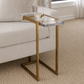 A modern side table with a clear acrylic tray top and textured gold metal frame, styled next to a light beige sofa on a woven rug.