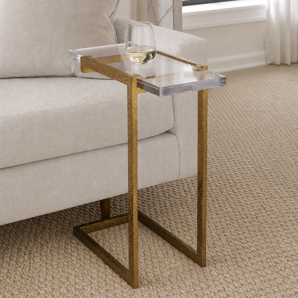 A modern side table with a clear acrylic tray top and textured gold metal frame, styled next to a light beige sofa on a woven rug.