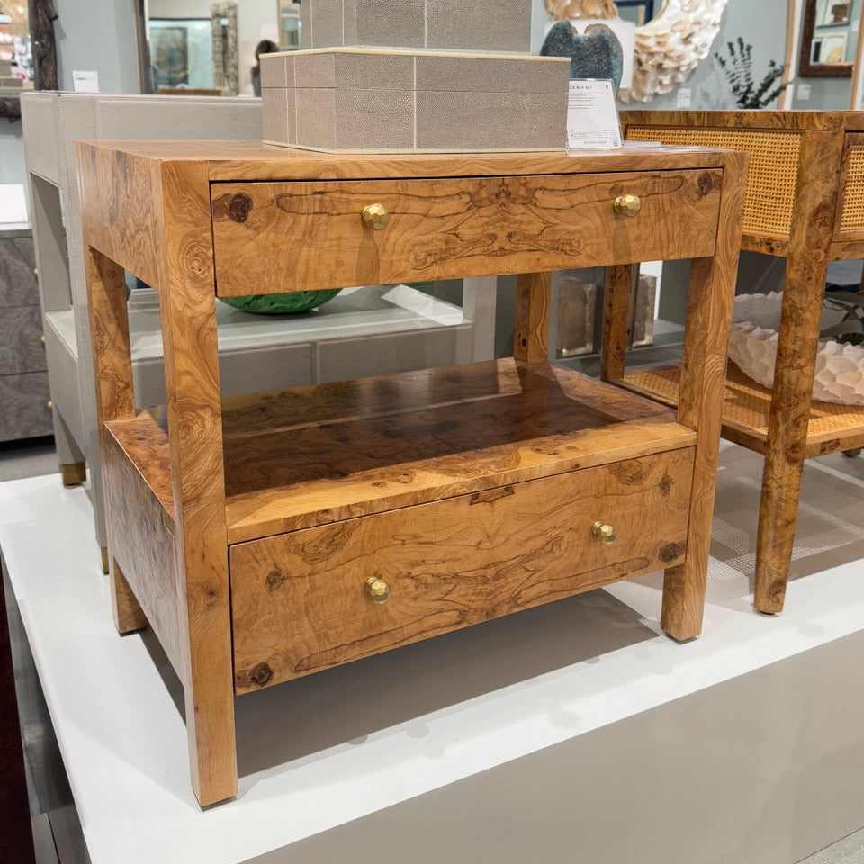 A wooden two-drawer console table with a natural burl wood finish and brass hardware, displayed on a white pedestal in a furniture showroom.