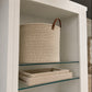 A large cylindrical woven storage basket in natural beige rope with a brown leather handle, displayed on a glass shelf inside a white cabinet. A matching woven tray sits on the lower shelf.