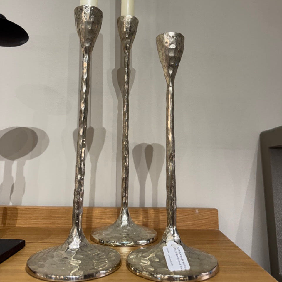 Three silver candle holders with candles on a wooden surface against a plain wall.