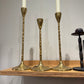 Three gold candle holders with white candles on a wooden shelf against a gray wall.