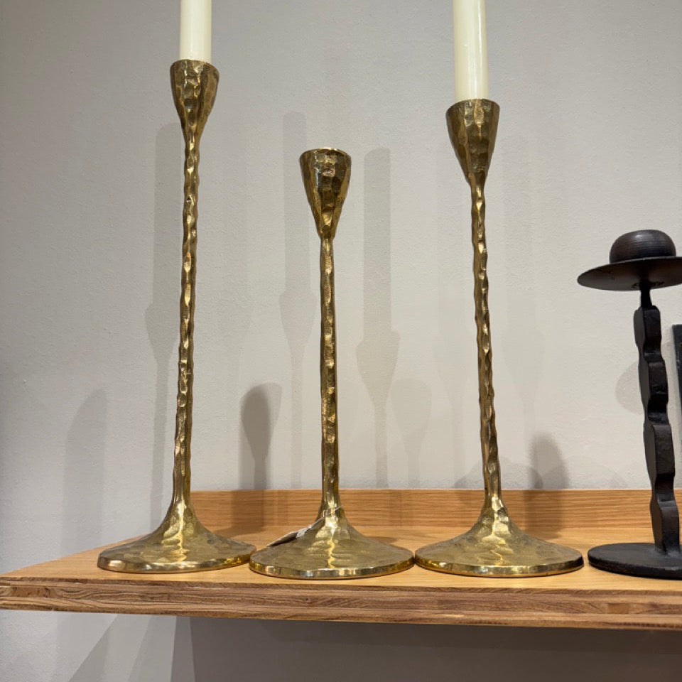 Three gold candle holders with white candles on a wooden shelf against a gray wall.