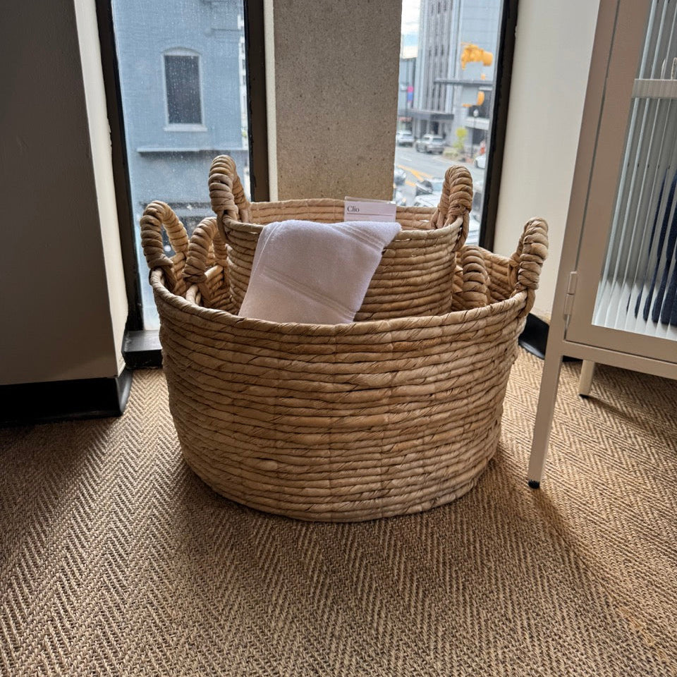 Woven basket with a white towel inside on a carpeted floor near a window.