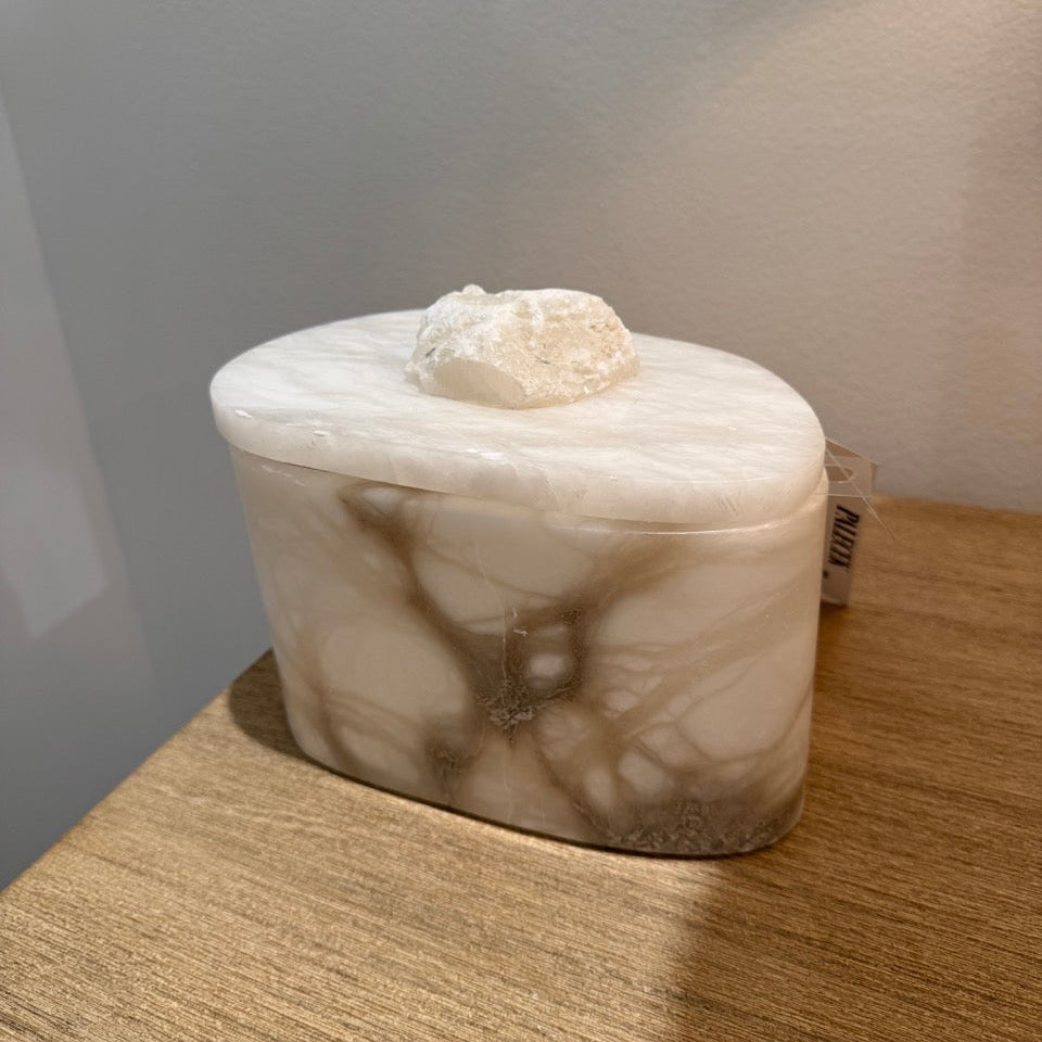 An oval-shaped resin box with a lid, featuring a white and beige marble-like pattern, sitting on a wooden tabletop.