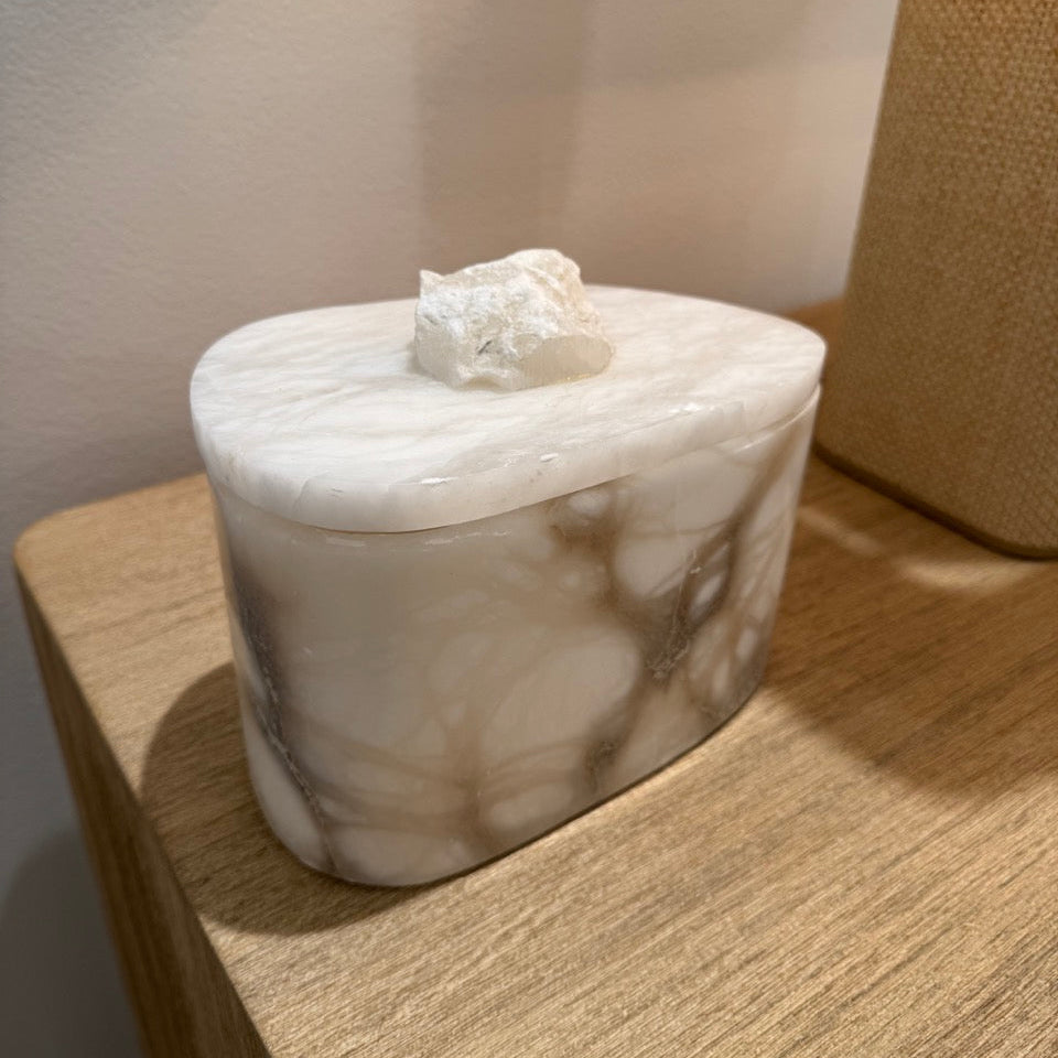 A small white marble tissue box cover or decorative box with a lid and rough stone-like top, shown sitting on a wooden table in an indoor setting.