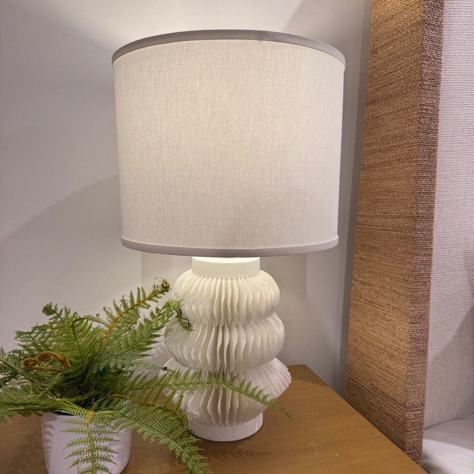 White textured table lamp with a beige lampshade on a wooden surface next to a plant.