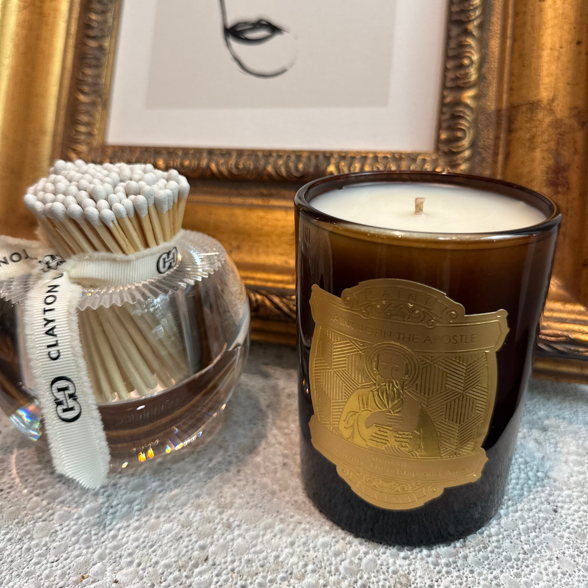 Candle with gold design, matches, and framed artwork on a textured surface