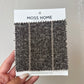 Moss Home fabric sample card held by a hand against a gray background