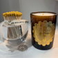 Candle with decorative label next to a glass holder with matches on a white surface.