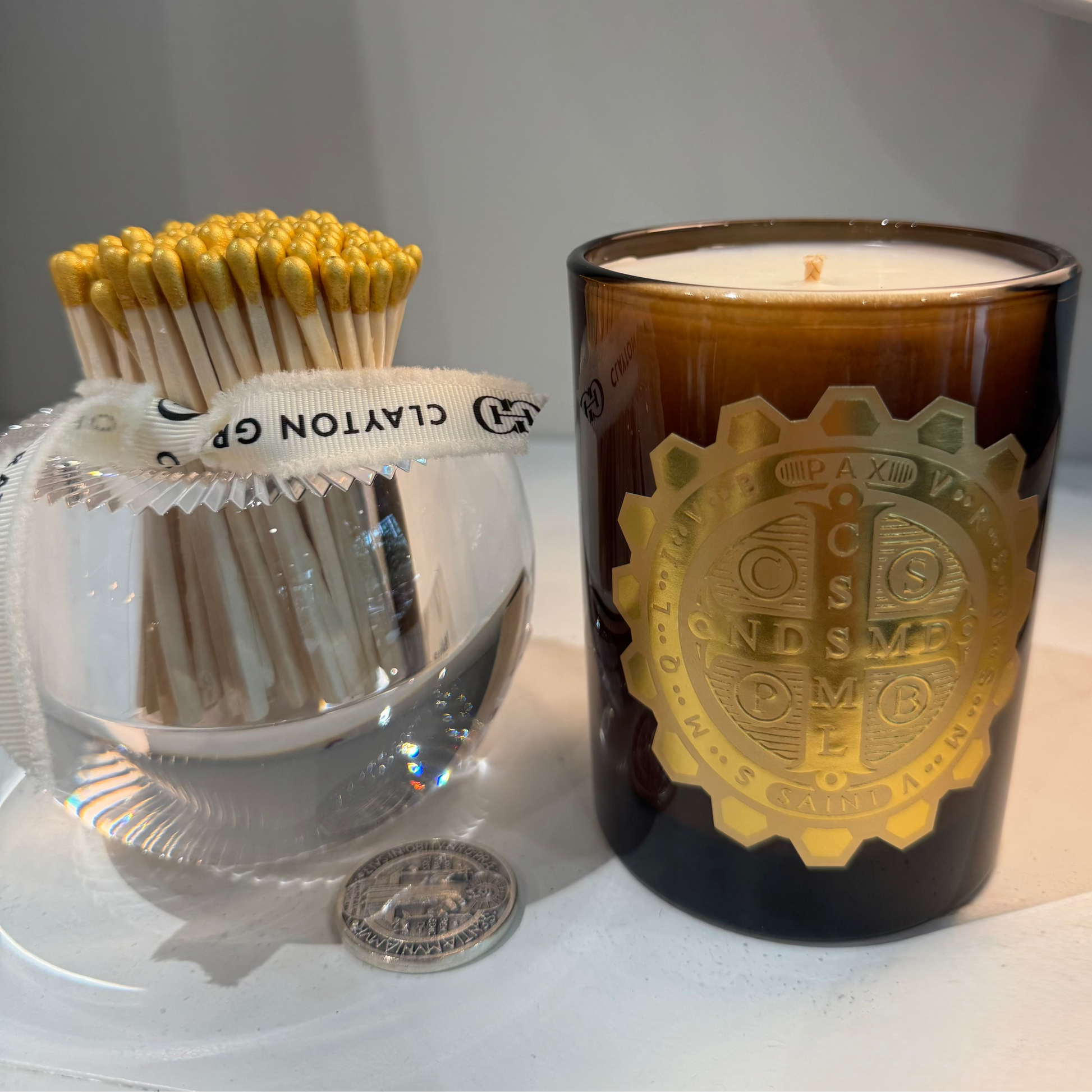 Candle with decorative label next to a glass holder with matches on a white surface.