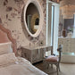 Neatly made bed in a bedroom with floral wallpaper, round mirror, and desk.
