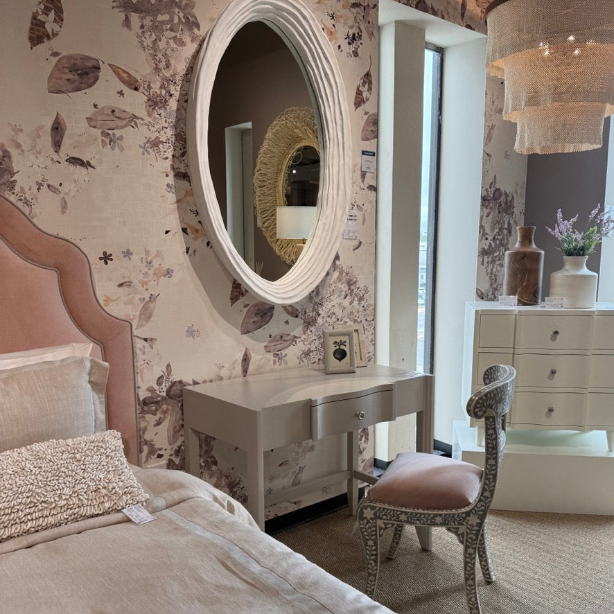 Neatly made bed in a bedroom with floral wallpaper, round mirror, and desk.