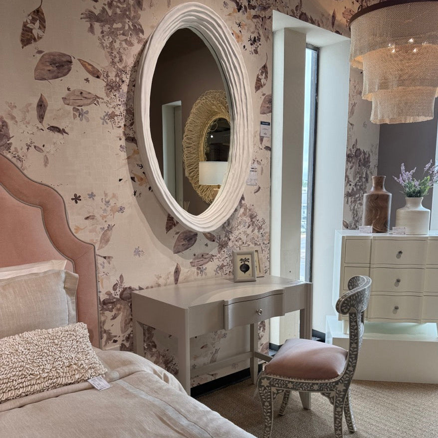 Neatly made bed in a bedroom with floral wallpaper, round mirror, and desk.