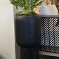 Tall black planter with a plant inside, placed on a surface with decorative elements in the background.