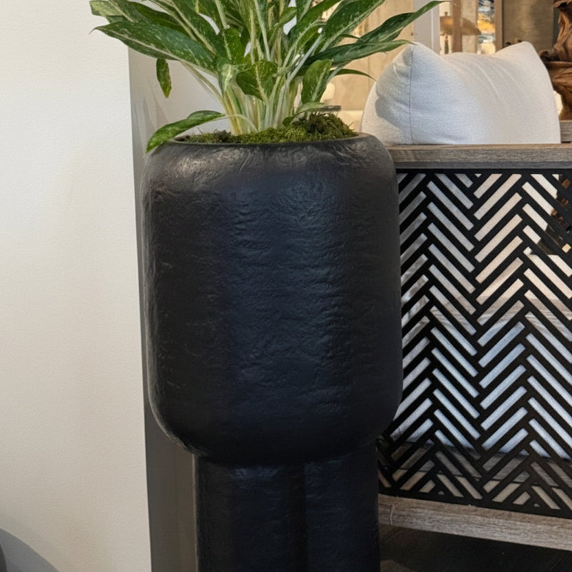 Tall black planter with a plant inside, placed on a surface with decorative elements in the background.