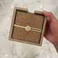 Brown textured jewelry box with a white tag held by a hand on a reflective surface.