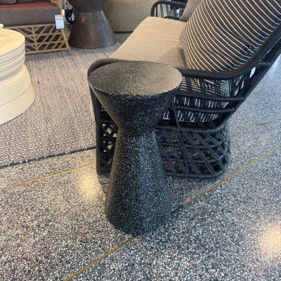 Black textured stool in front of a patterned chair on a speckled floor.