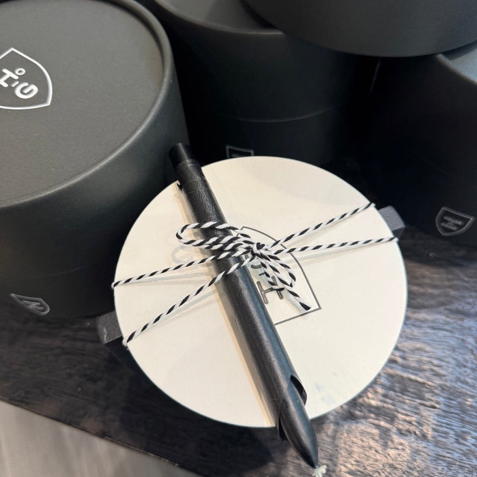 Black pen tied with string on a white circular card, surrounded by black cylindrical containers.