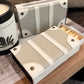 A close-up of a matchbox-style holder or case with a textured light gray and off-white leather-like finish, shown both closed and open with wooden matches visible. A candle jar and a book are partially visible in the styled setting.