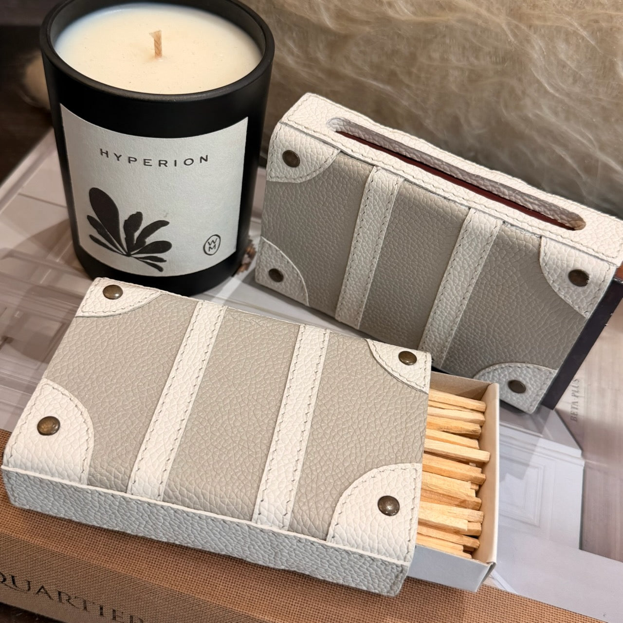 A set of two decorative matchbox-style candle holders in a light beige and white faux-leather material, shown with one filled with a black candle labeled HYPERION and the other open with wooden matches inside.