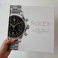 Hand holding a model of a Rolex watch against a white background with a Rolex promotional poster.
