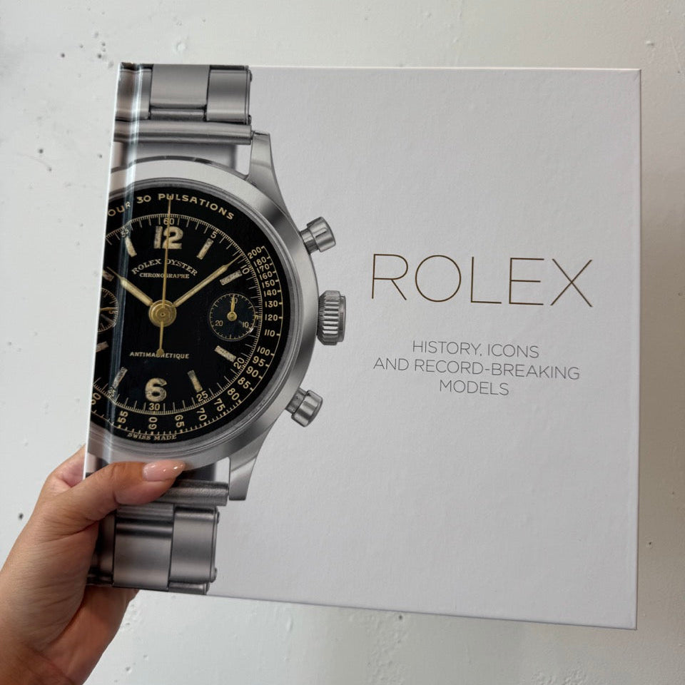 Hand holding a model of a Rolex watch against a white background with a Rolex promotional poster.
