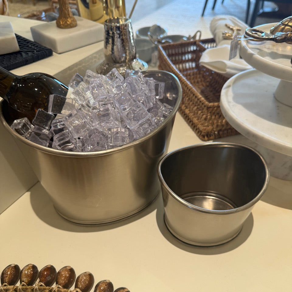 Metallic ice bucket with ice cubes and a small metal cup on a table.