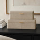 Two stacked rectangular storage boxes with a textured beige shagreen-like finish and slim gold handles, displayed on a dark wooden table in an interior setting.