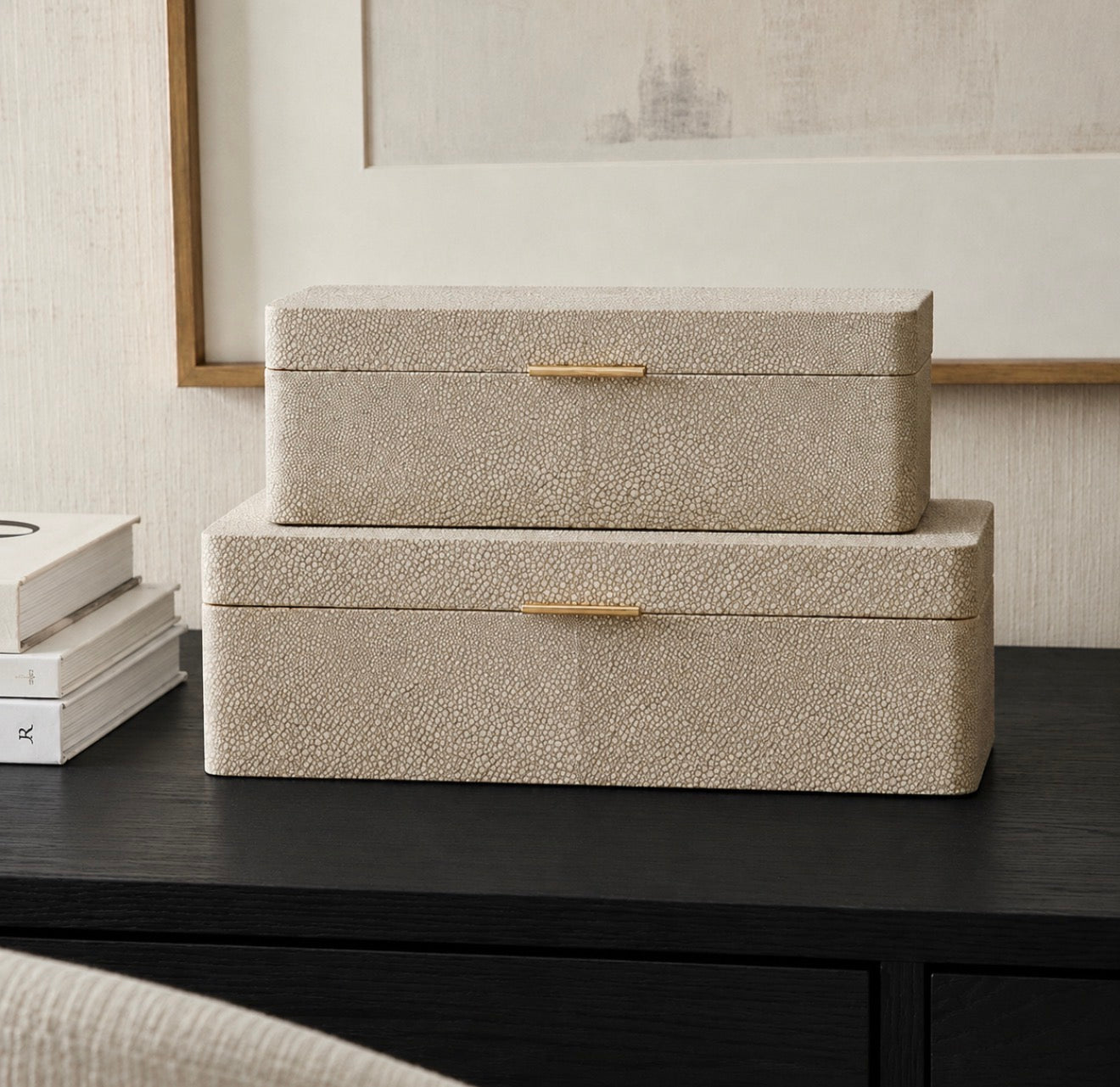 Two stacked rectangular storage boxes with a textured beige shagreen-like finish and slim gold handles, displayed on a dark wooden table in an interior setting.