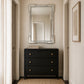 A black three-drawer dresser with brass knobs stands against a beige wall beneath a large rectangular mirror with a silver frame. The room has cream walls, side doors, a woven rug, and a framed print.