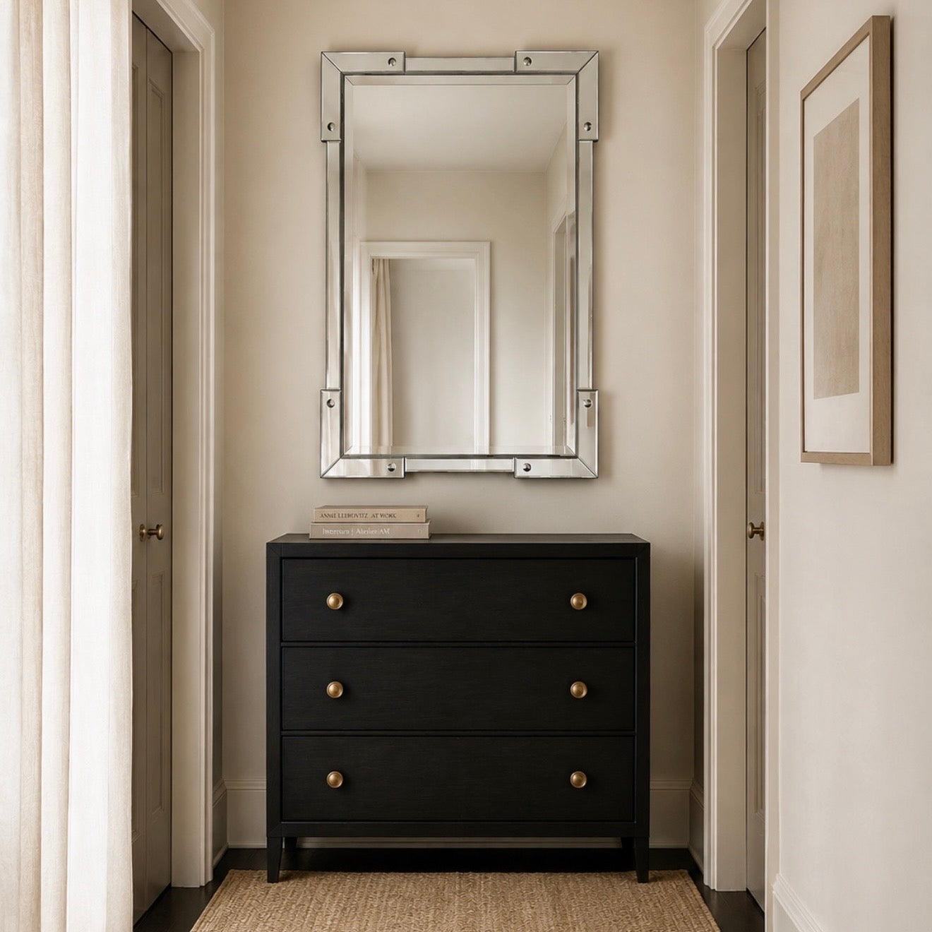 A black three-drawer dresser with brass knobs stands against a beige wall beneath a large rectangular mirror with a silver frame. The room has cream walls, side doors, a woven rug, and a framed print.