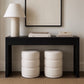 A black console table styled in a minimalist room setting with two cream-colored cylindrical ottomans underneath, a matching table lamp on the left, and books on top, against a neutral beige wall.
