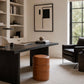 A modern home office scene featuring a dark wood writing desk as the primary product, styled with a brown leather barrel stool, a black desk lamp, and a black leather armchair in a bright, neutral room.