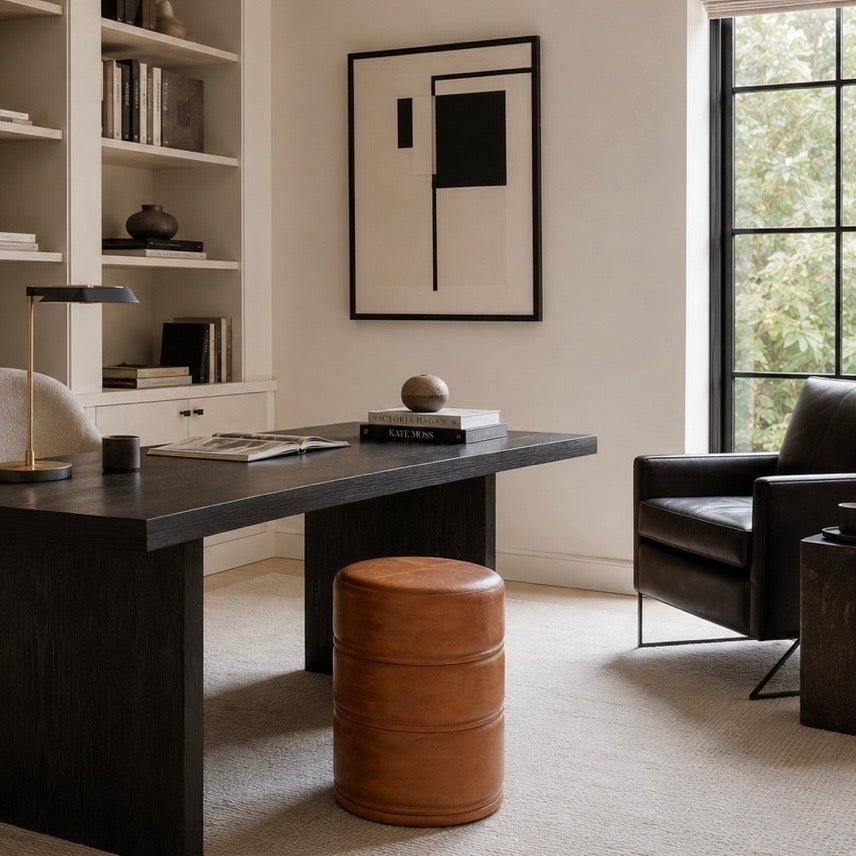 A modern home office scene featuring a dark wood writing desk as the primary product, styled with a brown leather barrel stool, a black desk lamp, and a black leather armchair in a bright, neutral room.