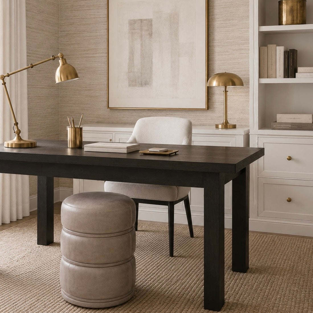 A dark wood writing desk in a styled home office setting, paired with a light beige upholstered chair, a round gray ottoman, a brass desk lamp, and a white built-in bookshelf in the background.