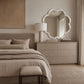 A large cloud-shaped mirror with a textured white frame hangs on a beige wall above a light wood dresser in a styled bedroom. The scene includes a bed with neutral bedding, a table lamp, and a rug.