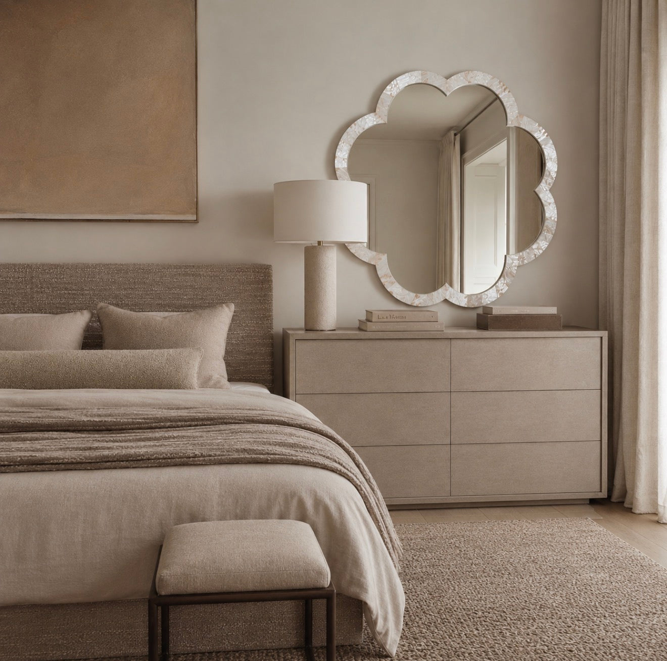 A large cloud-shaped mirror with a textured white frame hangs on a beige wall above a light wood dresser in a styled bedroom. The scene includes a bed with neutral bedding, a table lamp, and a rug.