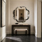 An ornate scalloped oval wall mirror with a dark marbled frame hangs above a dark wood console table and a light upholstered bench in a hallway with cream walls and dark stone flooring.