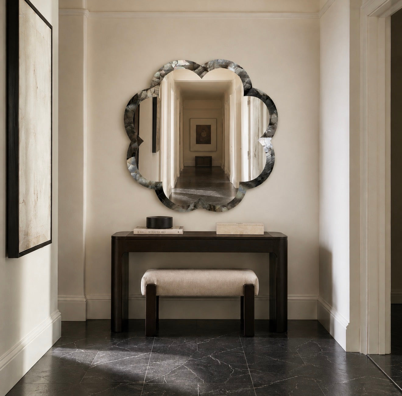 An ornate scalloped oval wall mirror with a dark marbled frame hangs above a dark wood console table and a light upholstered bench in a hallway with cream walls and dark stone flooring.