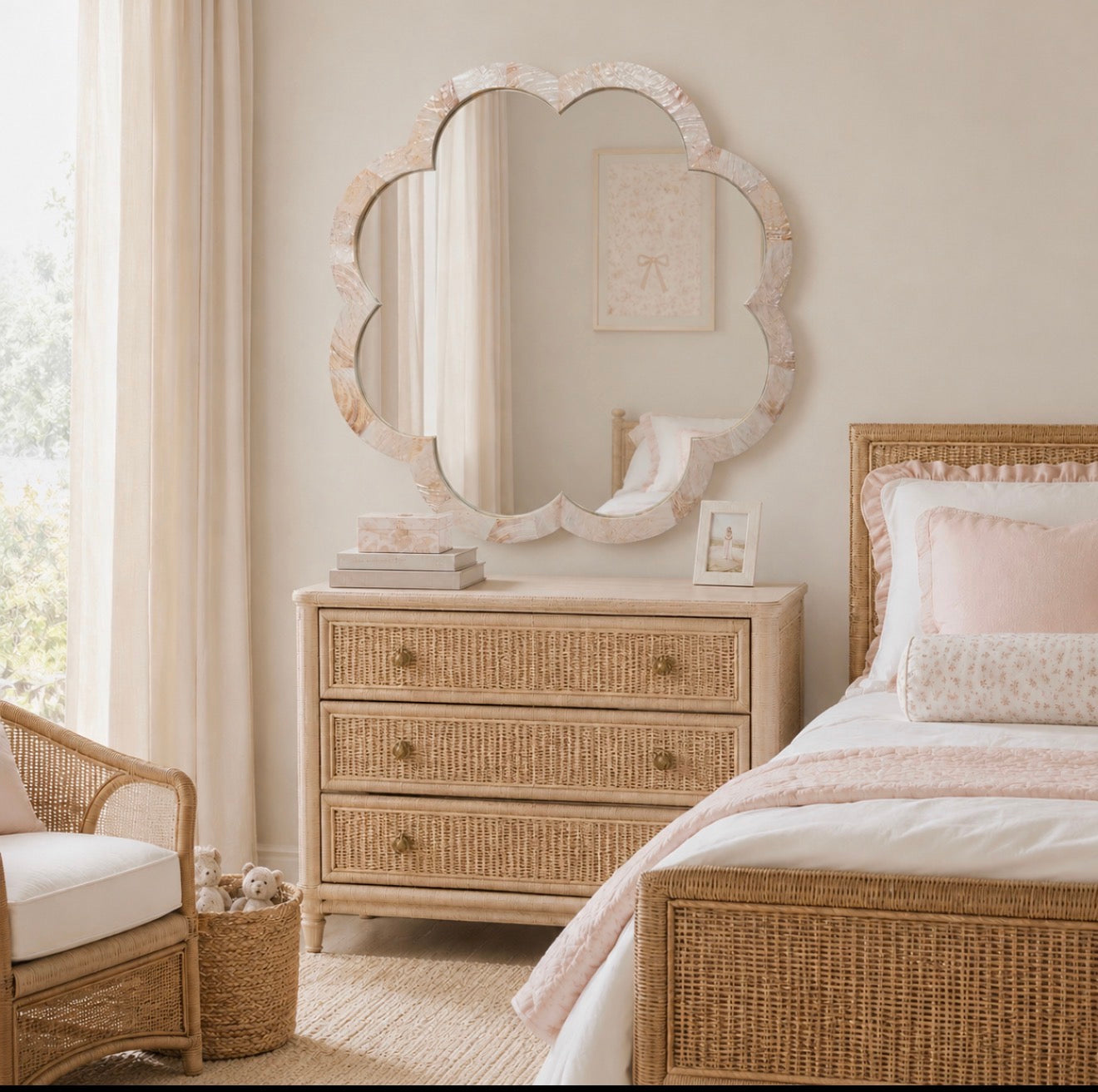 A styled bedroom scene featuring a large scalloped shell mirror with a distressed white and beige frame, mounted above a matching three-drawer rattan dresser. The room is decorated with a woven rattan bed frame, soft pink bedding, and a light beige armchair.