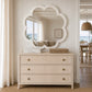 A coastal-style three-drawer dresser in a light beige woven rattan finish with brass knobs, styled under a matching scalloped cloud-shaped mirror in a bright, modern beachhouse interior.