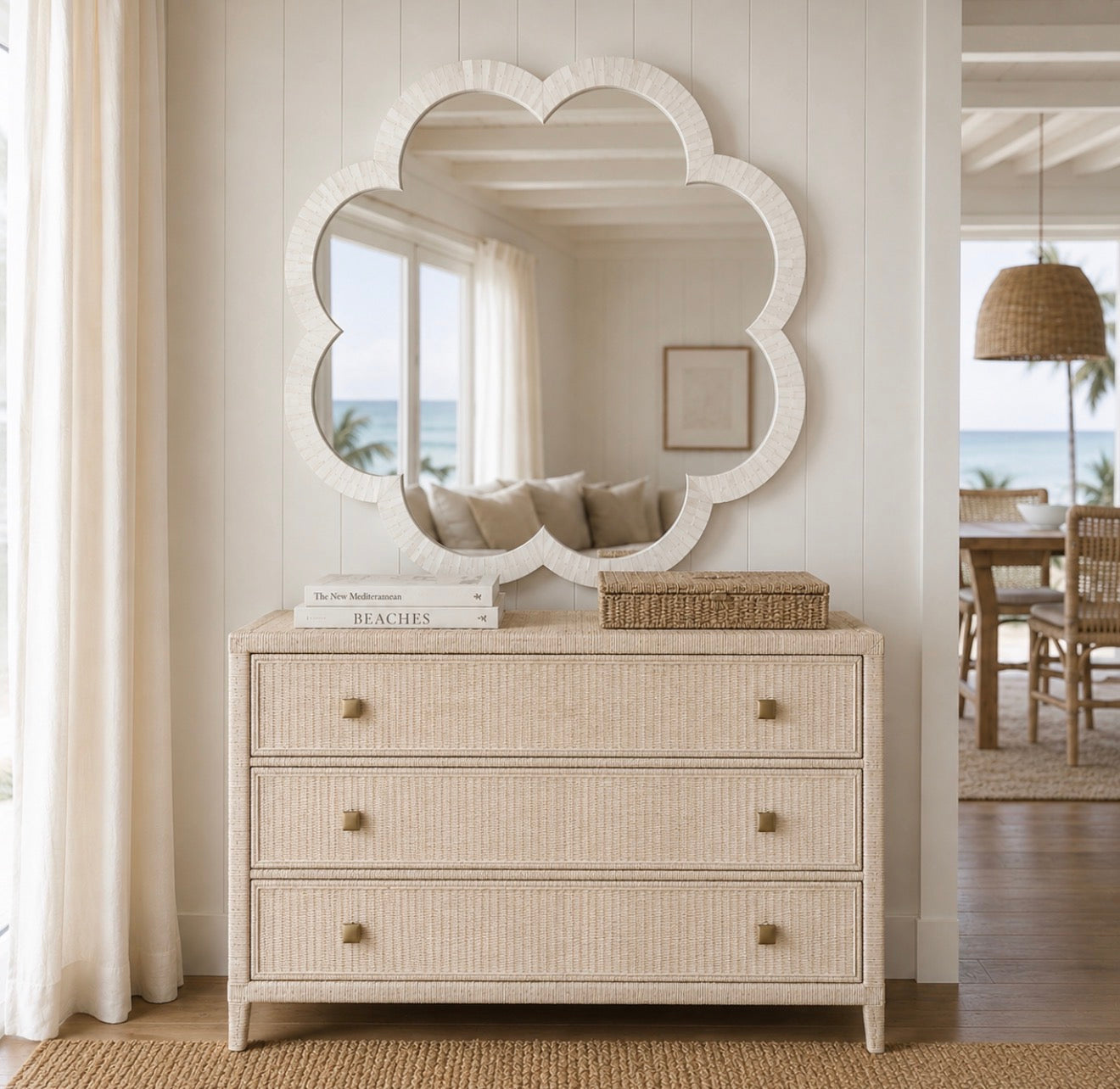 A coastal-style three-drawer dresser in a light beige woven rattan finish with brass knobs, styled under a matching scalloped cloud-shaped mirror in a bright, modern beachhouse interior.